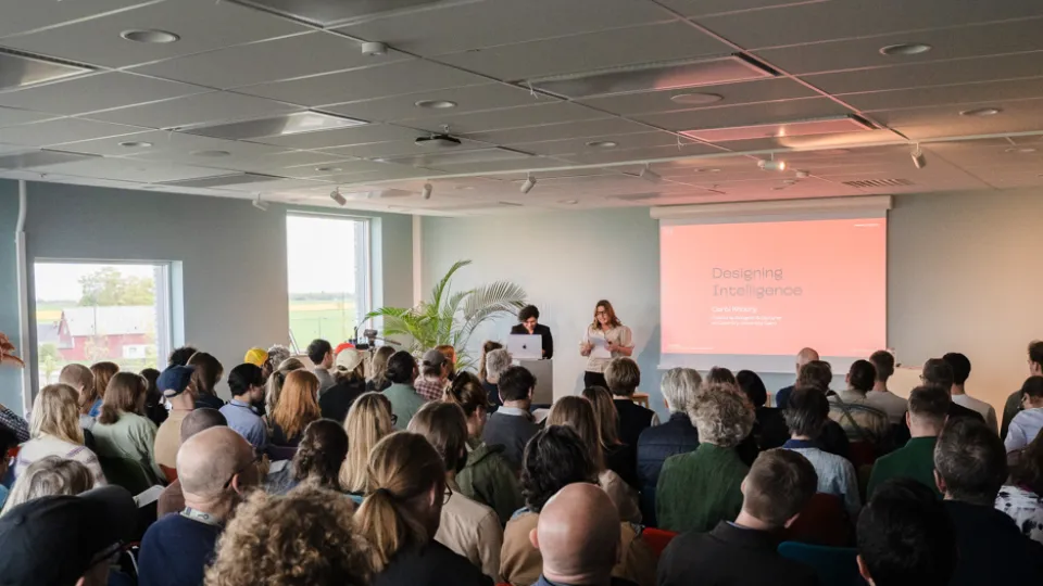 People seated in a large room facing two people standing up giving a presentation. Photo.