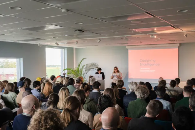 People seated in a large room facing two people standing up giving a presentation. Photo.
