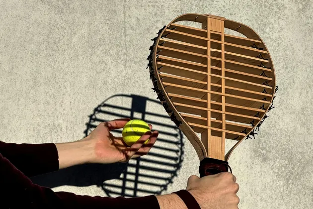 A wooden tennis racket casting a shadow on a tennis ball and the wall behind. Photo.