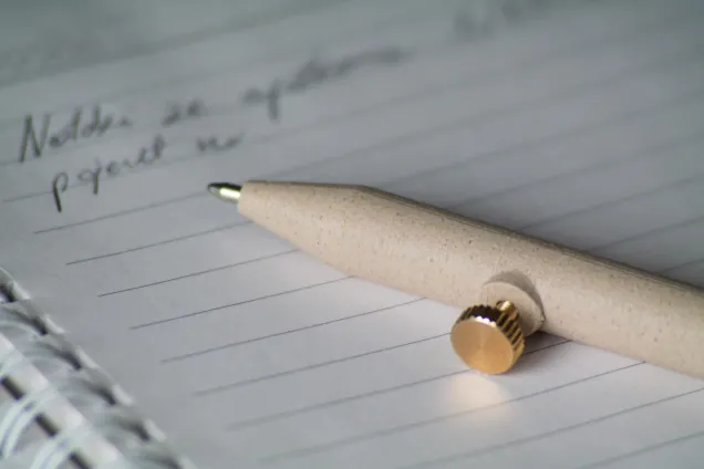 A beige pen with a gold button rests on an open lined notebook with faint handwritten text and visible spiral binding.