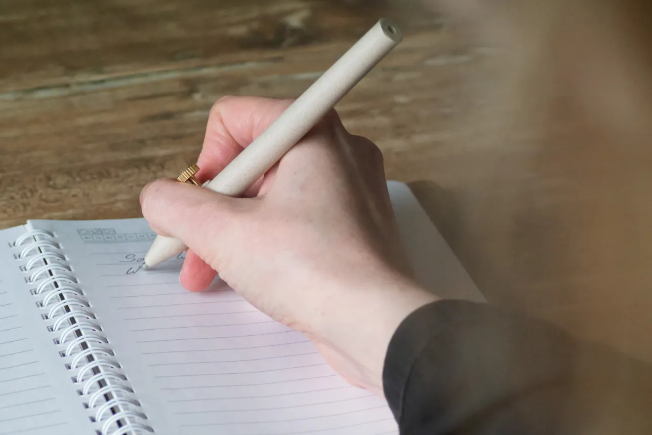 A hand holding a beige pen with a gold accent writes in a spiral-bound notebook on a wooden surface.