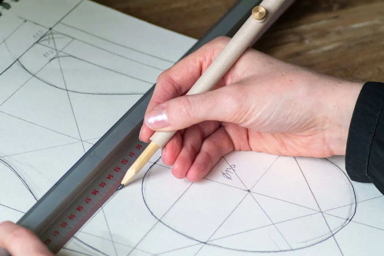 A hand uses a pencil and ruler to draw precise geometric shapes on technical drawing paper marked with circles, lines, and the 